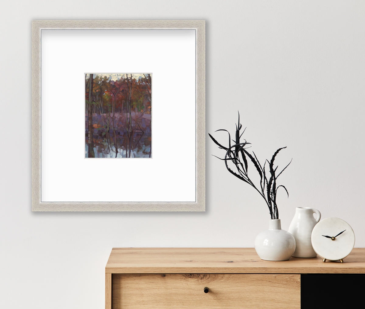 A muted multicolored abstract landscape print in a silver frame with a mat hangs on a white wall above a wood cabinet with a white modern clock, a small white pitcher and a white vase with dark green stems.