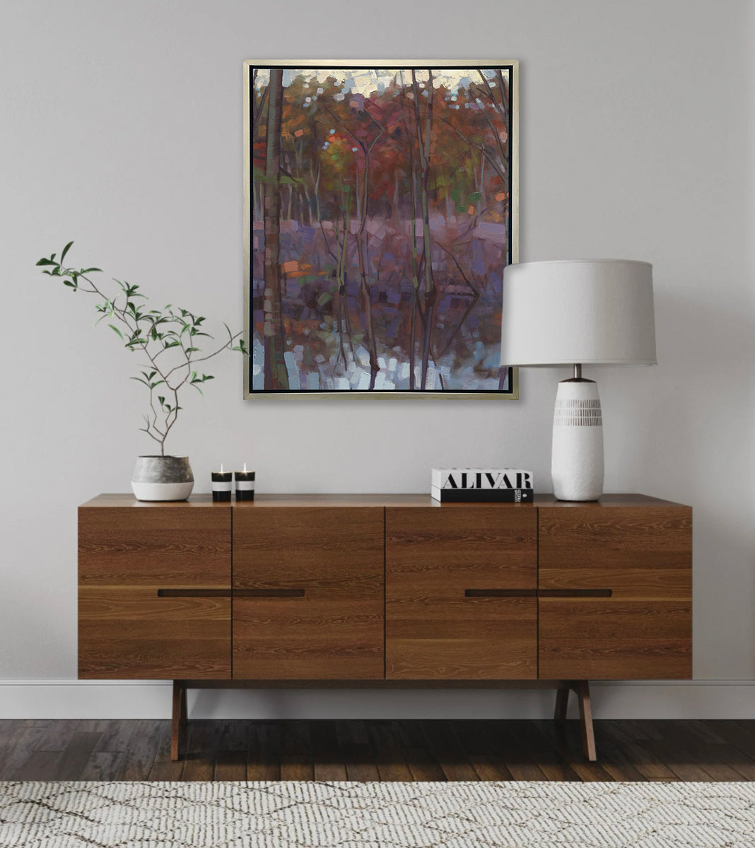 A muted multicolored abstract landscape print in a silver floater frame hangs on a white wall above a wood console cabinet with a white table lamp, a stack of books, two candles and a potted plant.
