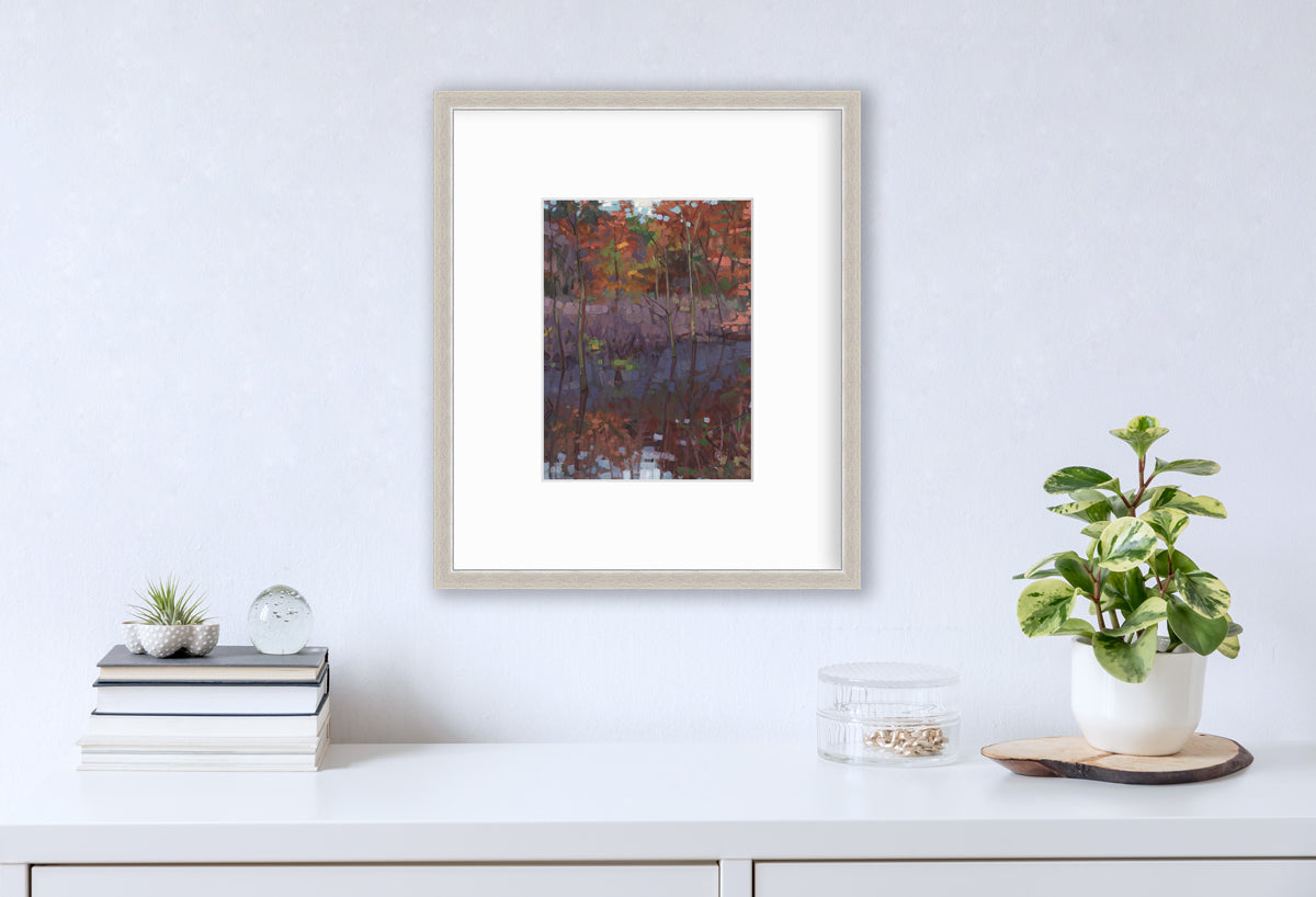 A muted multicolored abstract landscape print in a silver frame with a mat hangs on a white wall above a white surface with a stack of books, a round clear container with gold accessories and a potted plant.