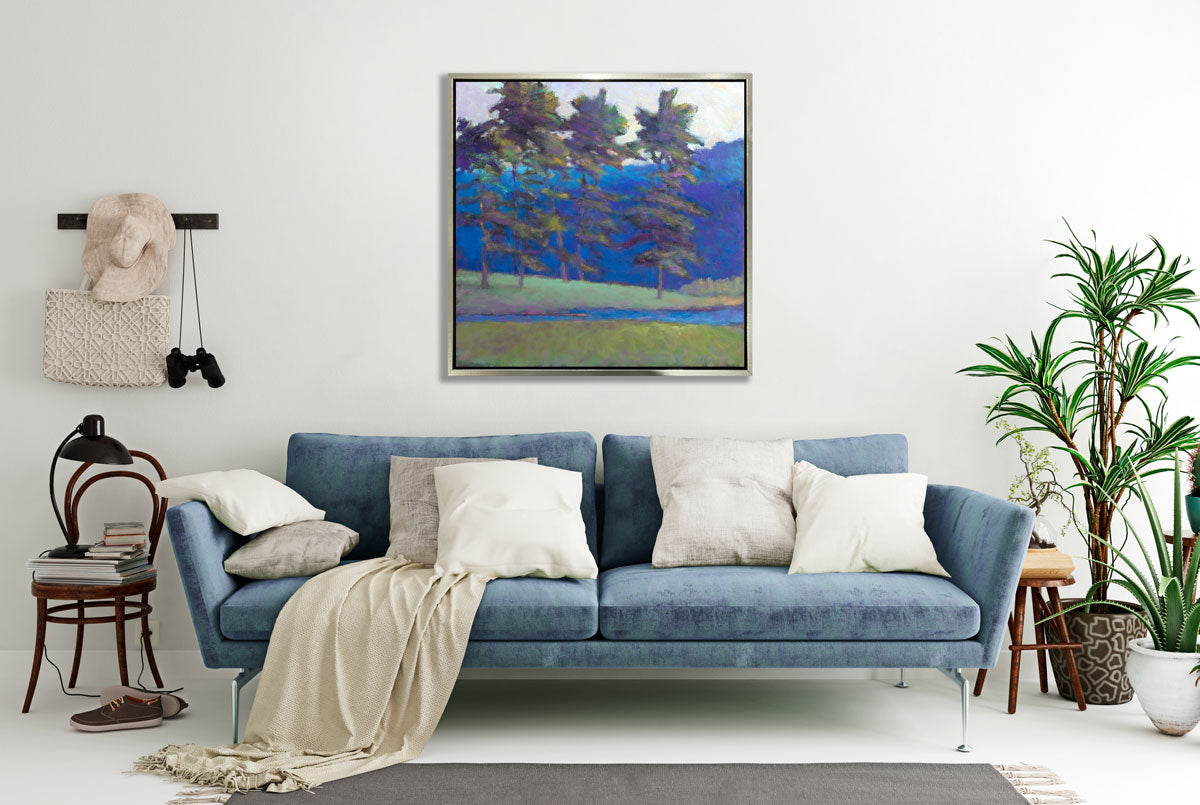 A blue and green tree landscape print in a silver floater frame hangs on a white wall above a blue sofa with white pillows and a blanket, two potted floor plants and a chair with books and a desk lamp.