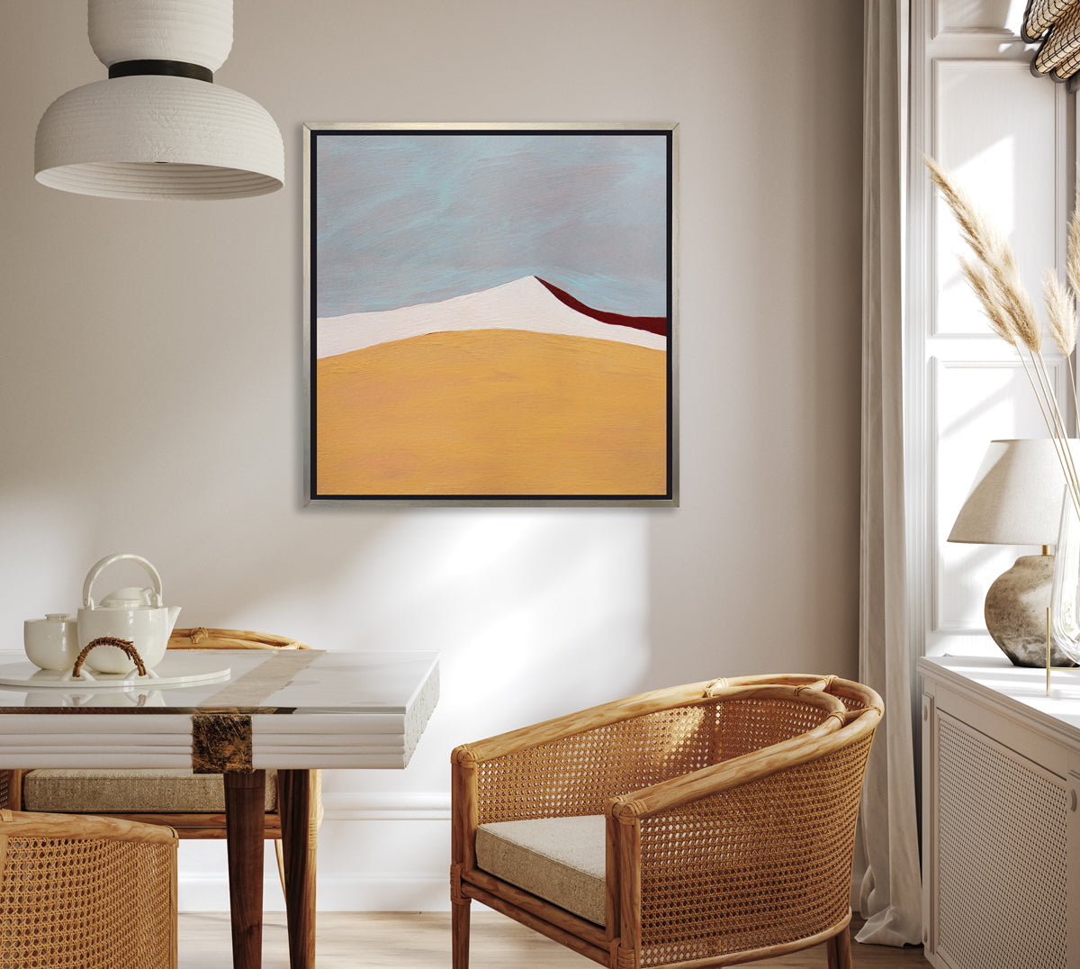 A dark yellow, white, dark red and slate blue abstract landscape print in a silver floater frame hangs on a white wall above a glass and wood table, natural rattan chairs and a window ledge with a table lamp.