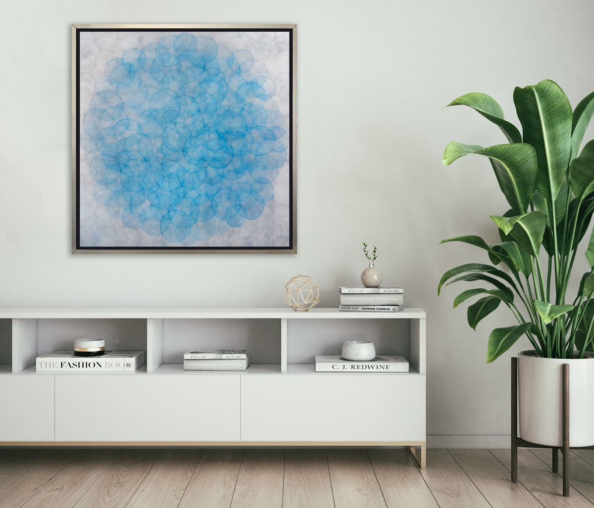 A silver and blue abstract geometric print in a silver floater frame hangs on a white wall above a white shelf credenza with stacks of books, a gold wire sphere decoration and a potted floor plant.