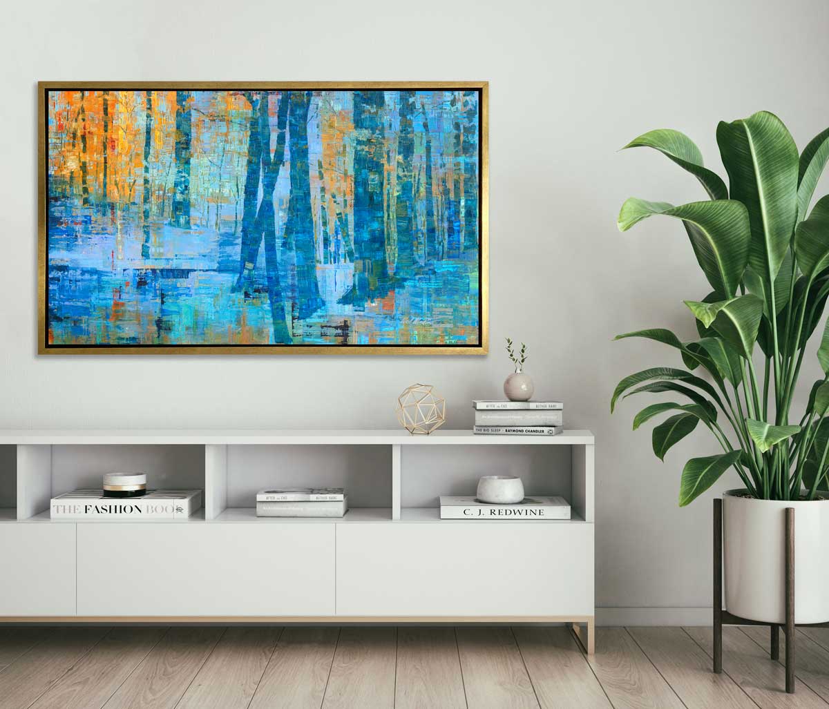 A blue, green and orange abstract tree landscape print in a gold floater frame hangs on a white wall above a white credenza with books, decorative accessories and a potted floor plant is to the right.
