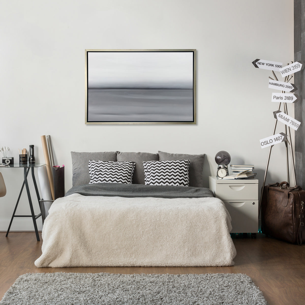 A grey toned abstract landscape print in a silver floater frame hangs on a white wall above a bed with dark grey bedding, a cream colored throw blanket and a small side table with decorative accessories.