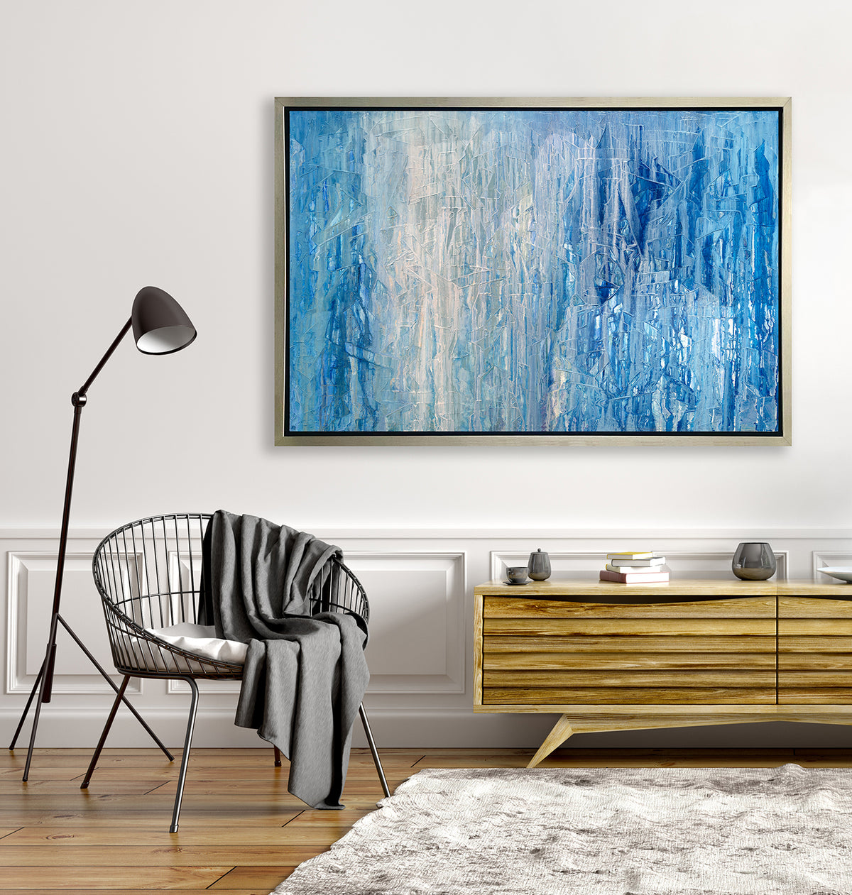 A blue, white and grey abstract print in a silver floater frame hangs on a white wall above a wooden console table with vases and a grey wire chair.