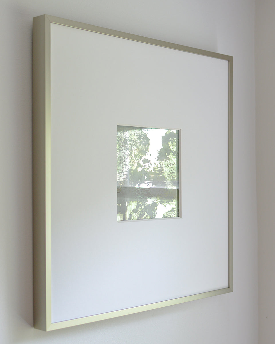 A left side-angle view of a small grey and silver painted art mirror in a silver frame with a large white mat hangs on a white wall.