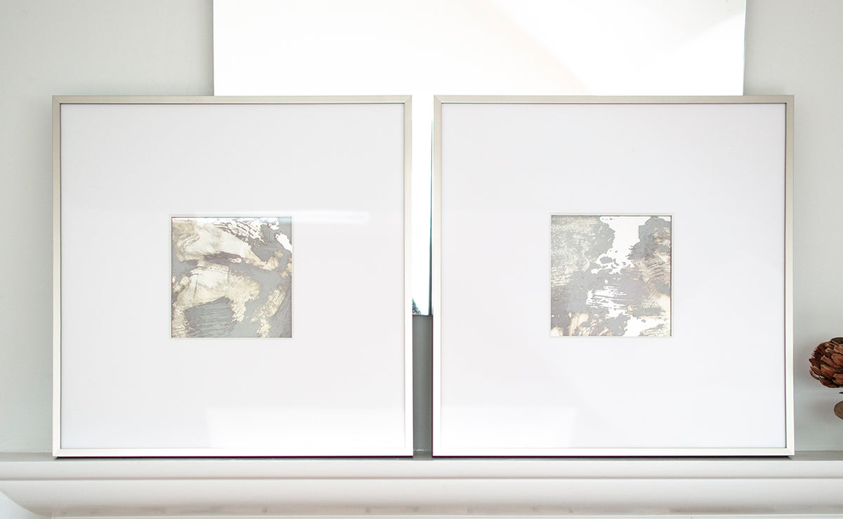 Two small grey and silver painted art mirrors in silver frames with large white mats sit on a white surface.