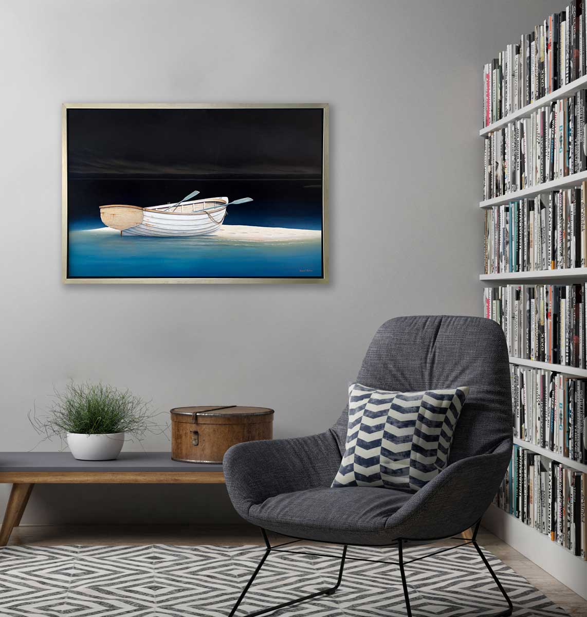 A contemporary realist print of a white rowboat alone on a sandbank in dark water against a dark sky in a silver floater frame hangs on a grey wall above a grey bench with a potted plant and a hat box, a grey chair with a grey and white printed pillow and a full wall bookshelf.