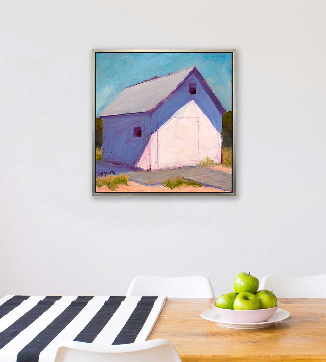A blue, white and lavender contemporary barn print in a silver floater frame hangs on a white wall above a wood table with a blue and white table runner and a bowl of green apples.