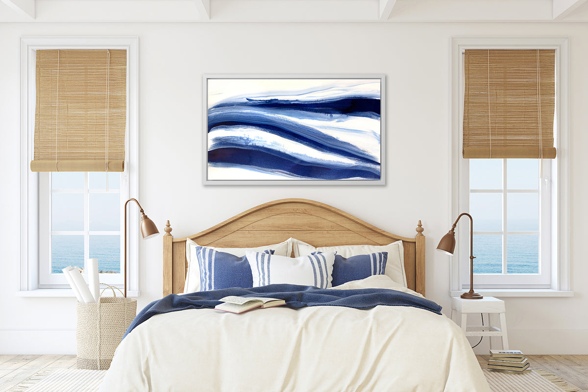 A dark blue and white abstract print in a white floater frame hangs on a white wall above a bed with a wood arched headboard, white bedding and blue and white striped pillows.