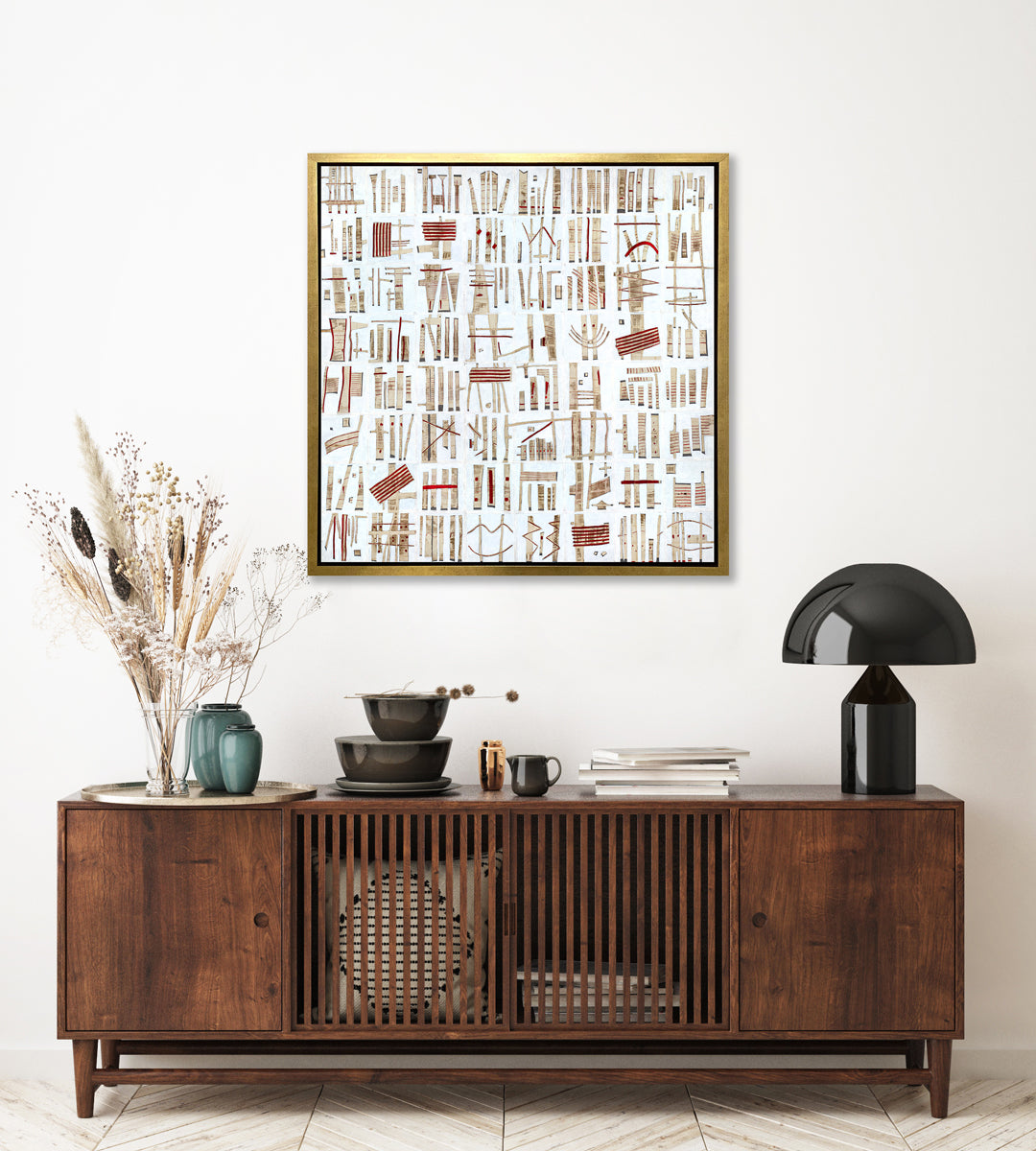 A white, beige and red abstract print in a gold floater frame hangs on a white wall above a dark wood cabinet with a black table lamp, a stack of brown bowls, a stack of books and a tray with two teal vases and a clear vase with wheat stems.