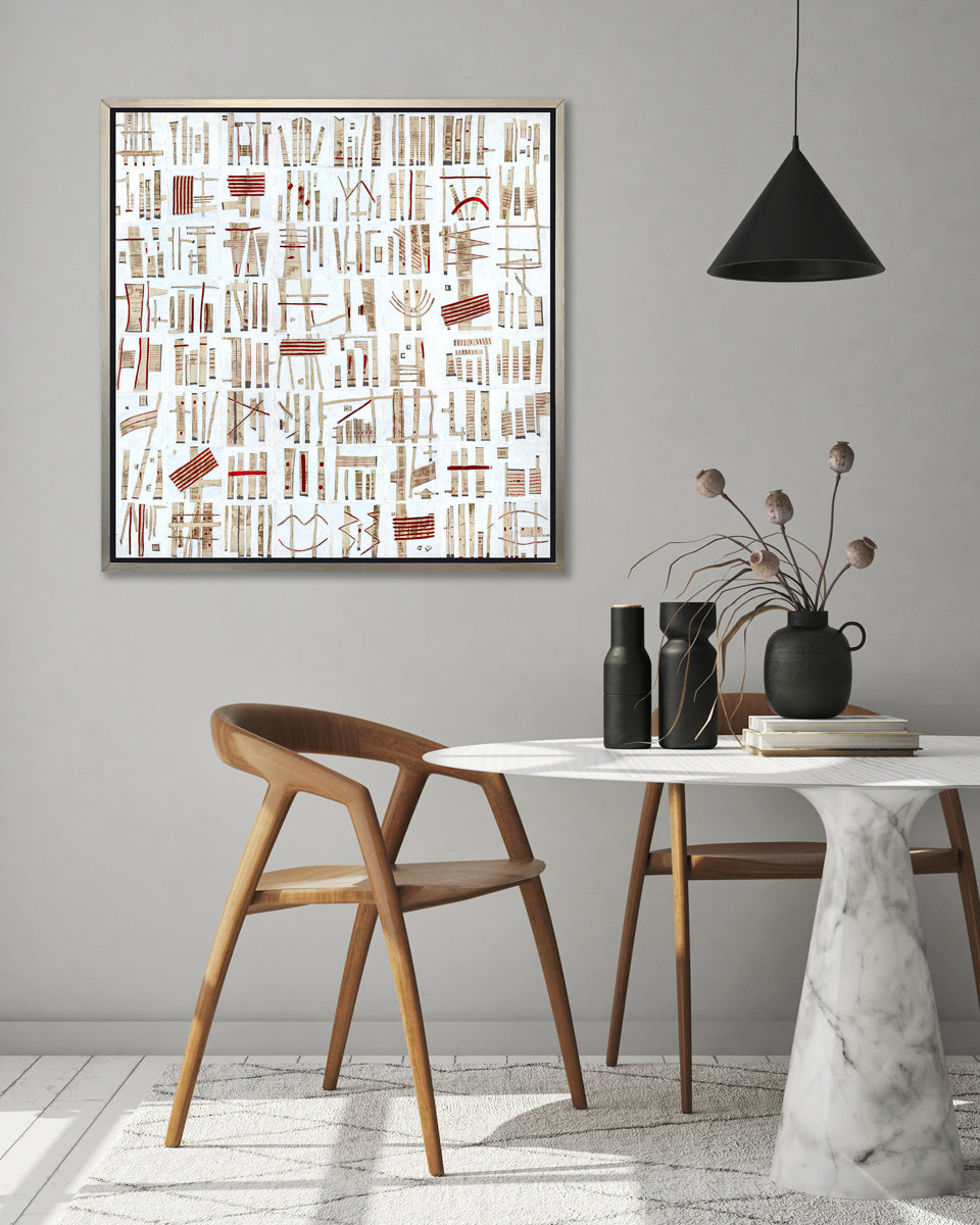 A white, beige and red abstract print in a silver floater frame hangs on a white wall above a round white marble table with black vases and two dark modern chairs.