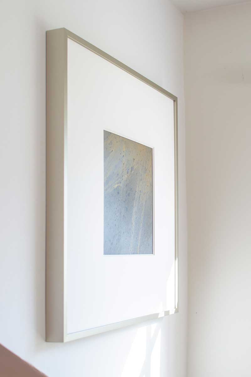 A left side-angle view of a small light yellow, light blue and silver painted art mirror in a silver frame with a large white mat hangs on a white wall.