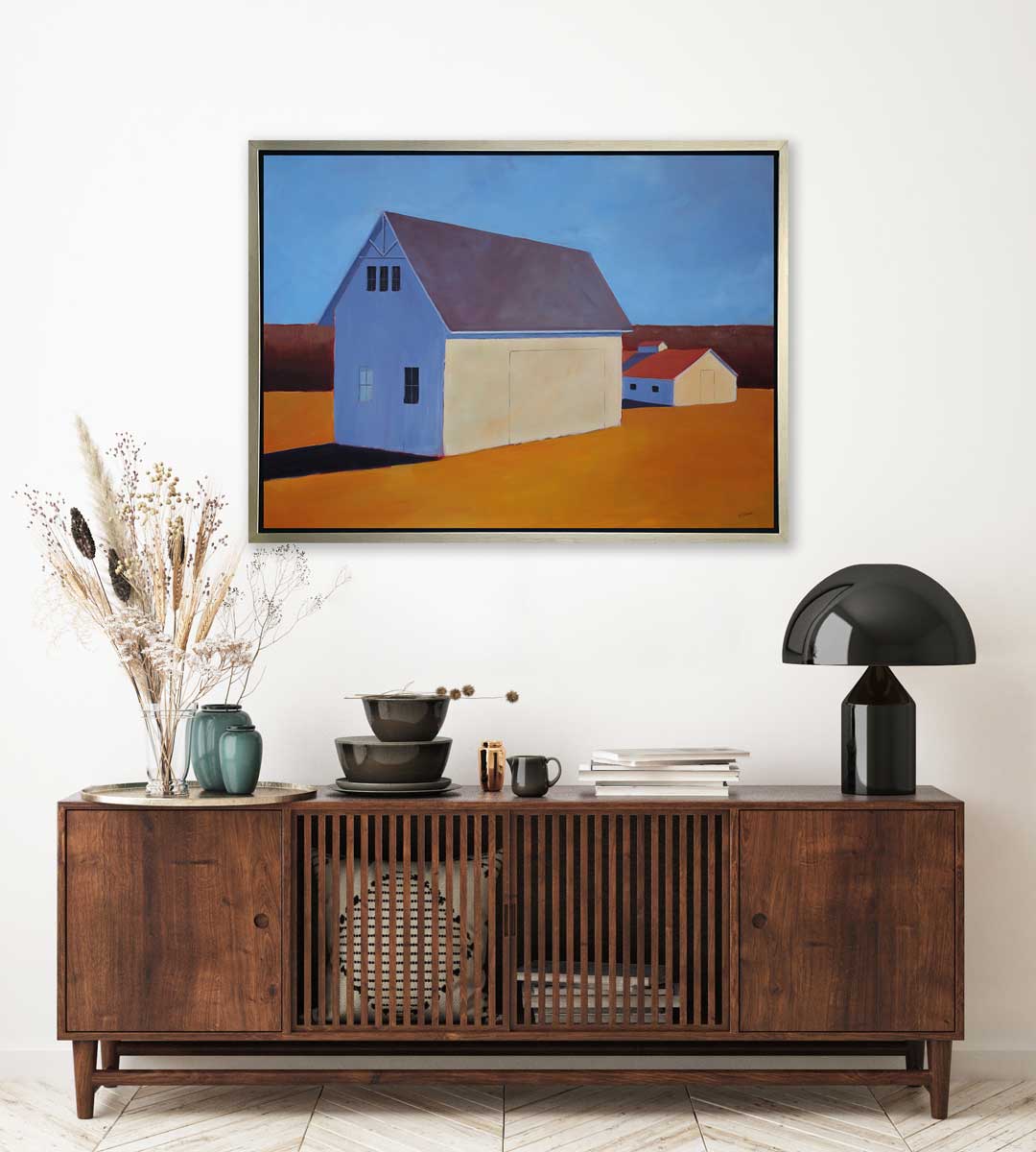 A blue, dark orange, beige and lavender contemporary barn print in a silver floater frame hangs on a white wall above a dark wood sideboard with a black modern lamp, a stack of books, a stack of brown dishes and a tray with a clear vase with stems.