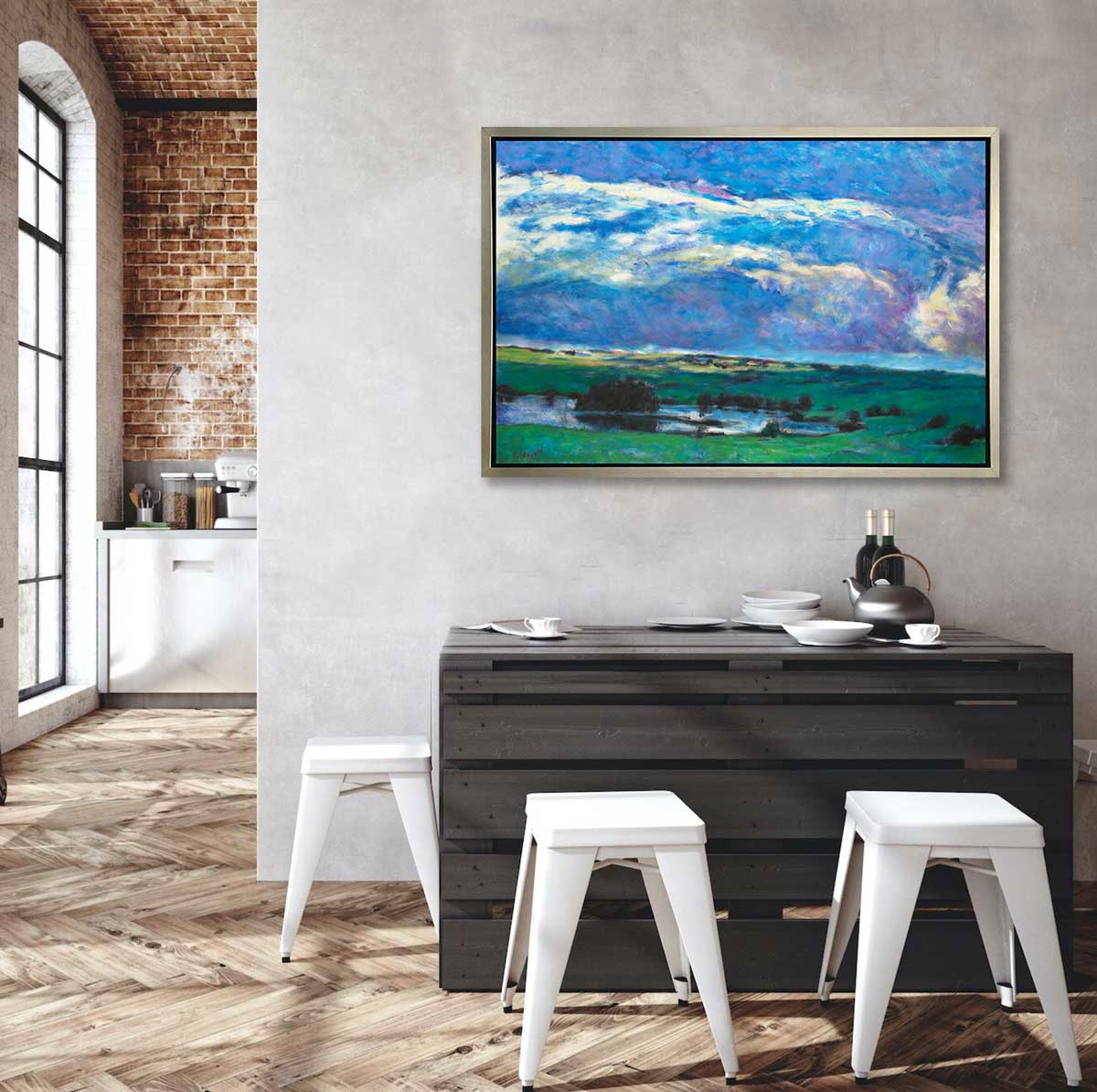 A blue and green abstract landscape print in a silver floater frame hangs on a grey wall above a black wood palette table with surrounding white backless stools.