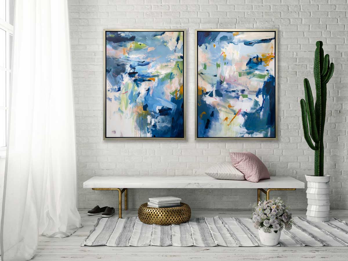 Two blue, green and white abstract prints in silver floater frames hang on a white brick wall above a white marble bench with pillows, a potted cacti and a gold decorative ottoman.
