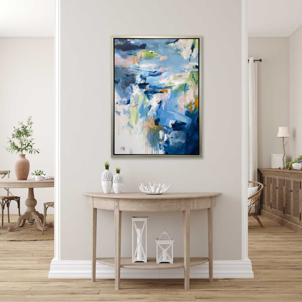 A blue, green and white abstract print by Kelly Rossetti in a silver floater frame hangs on a white wall above a natural wood half-circle console table with two white vases, a white decorative bowl and two white lanterns.