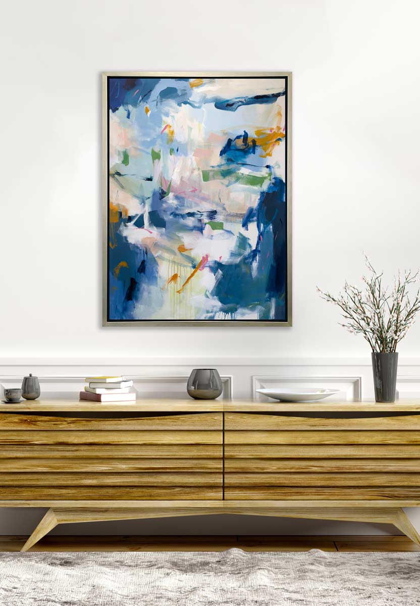 A blue, green and white abstract print in a silver floater frame hangs on a white wall above a wood console cabinet with chrome vases, a stack of books, a white plate and a grey vase with pink cherry blossom stems.