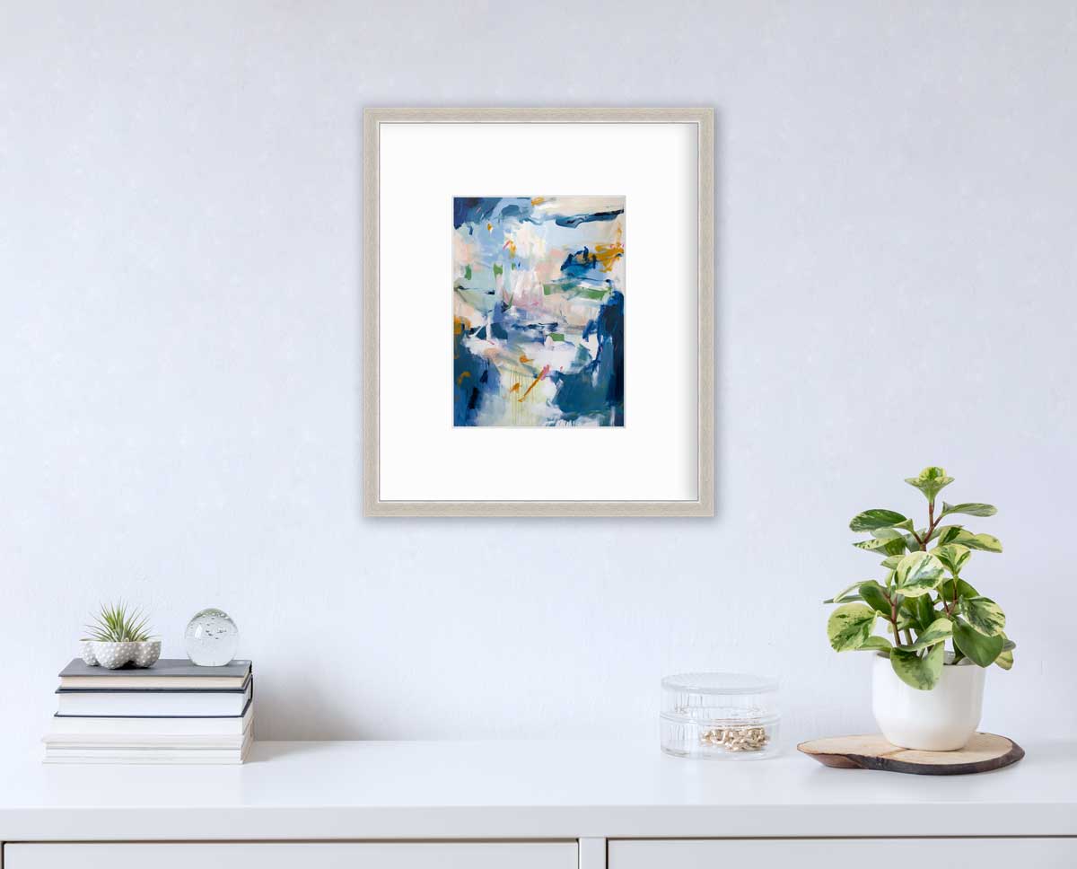 A blue abstract print in a silver frame with a mat hangs on a white wall above a white surface with a stack of books, a clear round container with gold accessories and a potted plant.