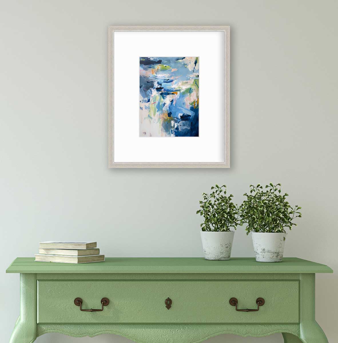 A blue abstract print by Kelly Rossetti in a silver frame with a mat hangs on a white wall above a green console table with a stack of books and two potted plants.