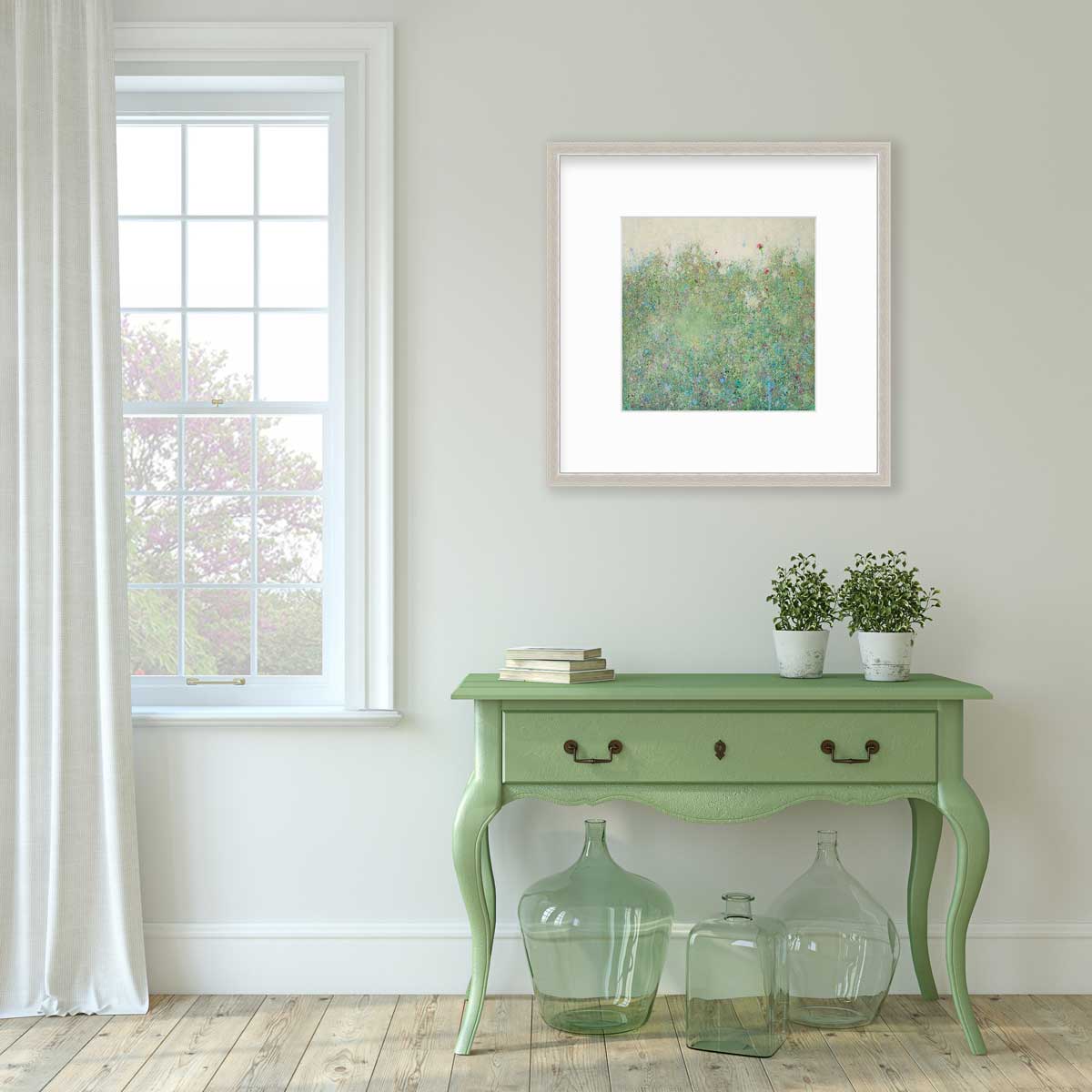 A green abstract landscape print in a silver frame with a mat hangs on a white wall above a green console table with a stack of books, two potted plants and three green glass bottles.