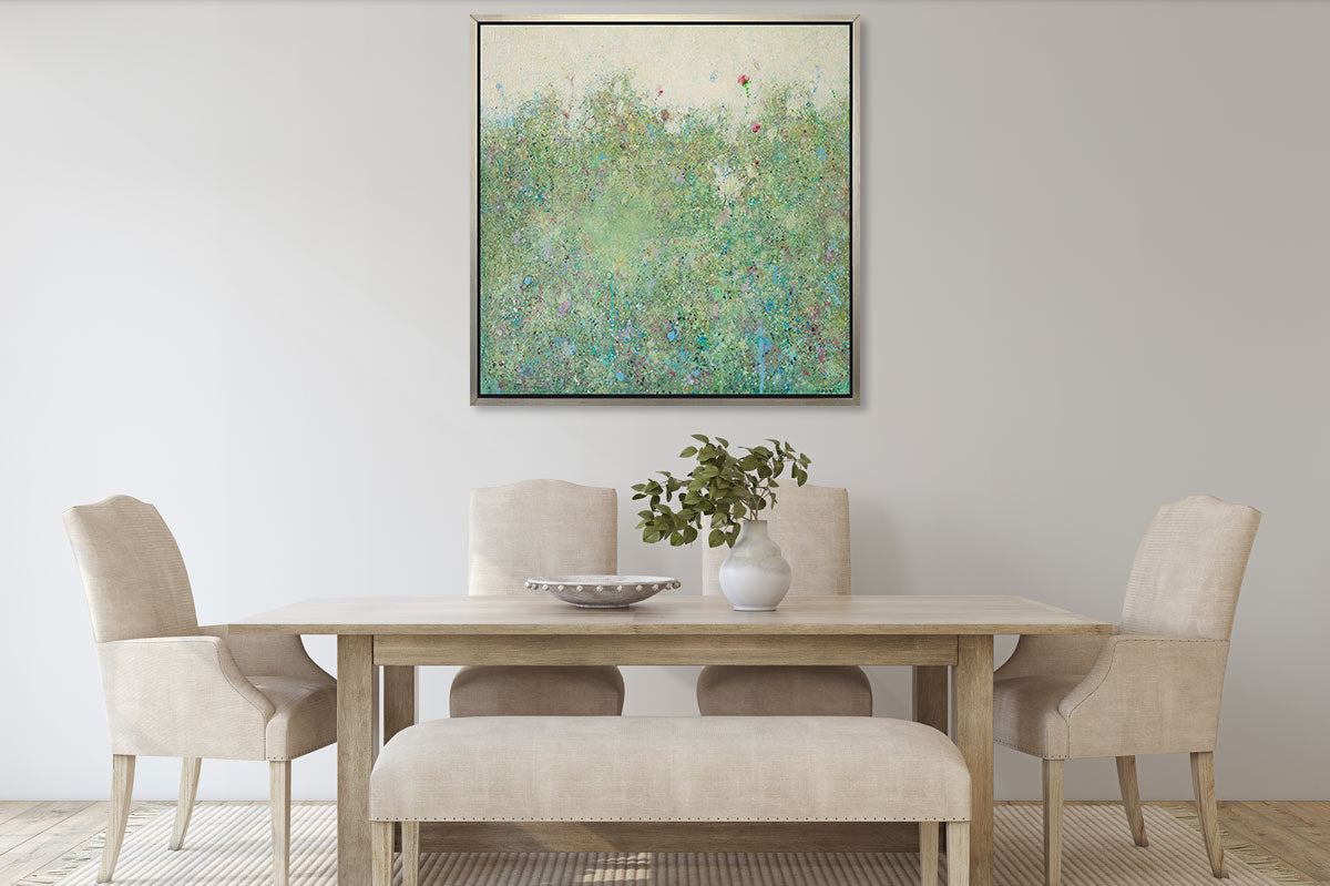 A green abstract landscape in a silver floater frame hangs on a white wall above a natural wood dining table, four beige cloth chairs and beige cushioned bench.