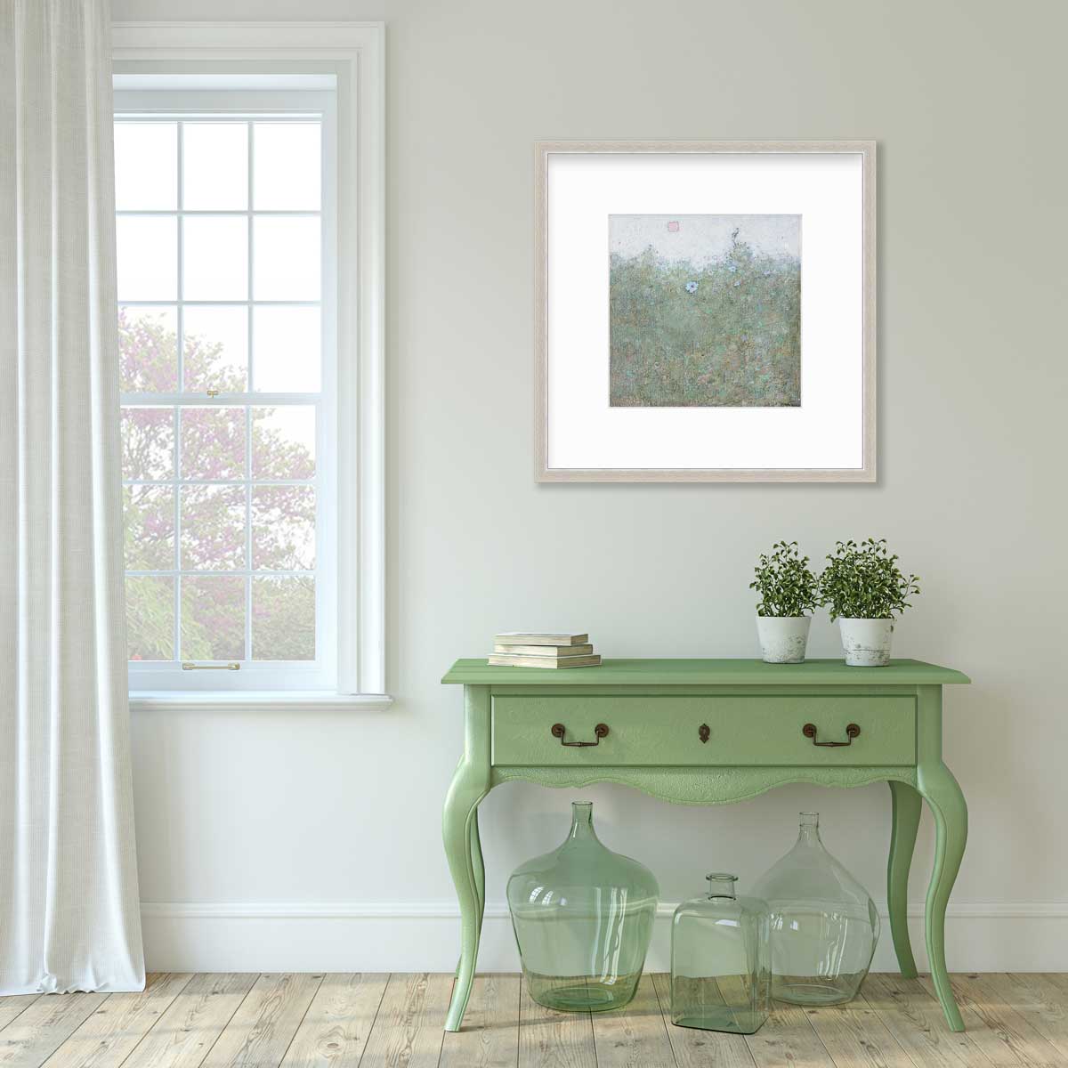 A green and blue abstract landscape print in a silver frame with a mat hangs on a white wall above a green console table with a stack of books, two potted plants and three green glass bottles.