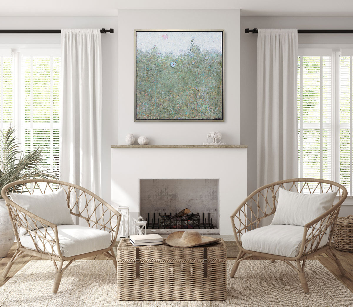 A blue and green abstract landscape in a silver floater frame hangs on a white wall above a fireplace, two rattan chairs with white cushions and a woven trunk.
