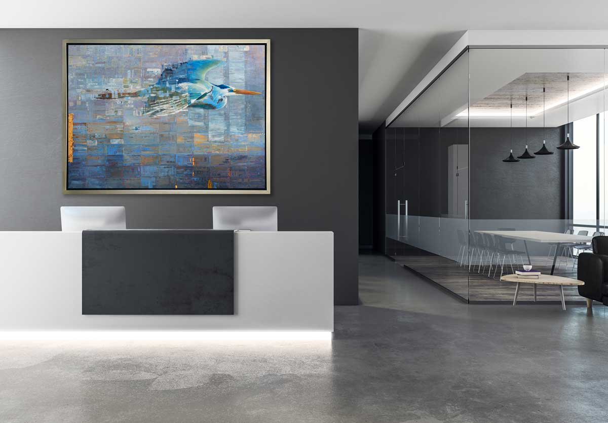 A blue, teal and light purple geometric abstract print of a blue heron in mid-flight with gold accents in a silver floater frame hangs on a dark grey wall above a large white desk with two computer monitors, across from a glass enclosed office with a table and chairs.