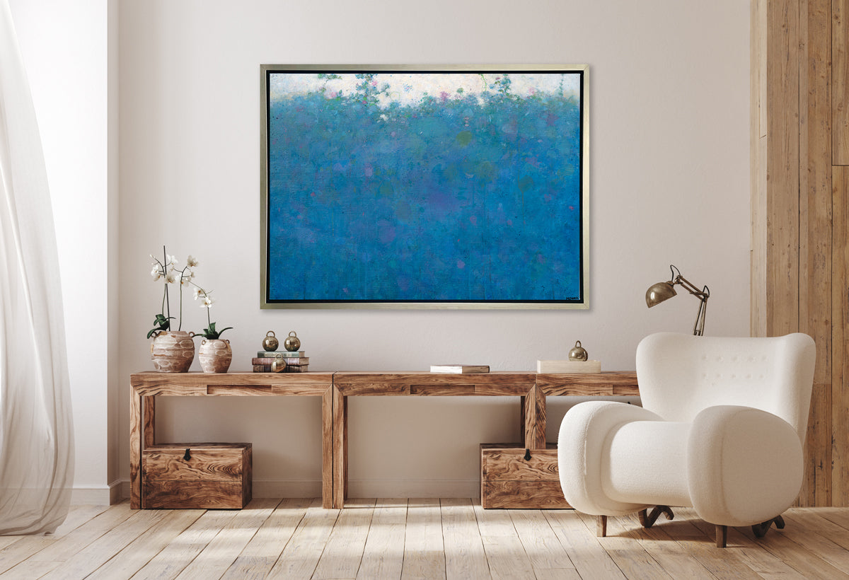 A blue, green, pink and white abstract landscape print in a silver floater frame hangs on a white wall above a long wooden desk with a stack of books, two potted white orchids and a white modern chair to the right.
