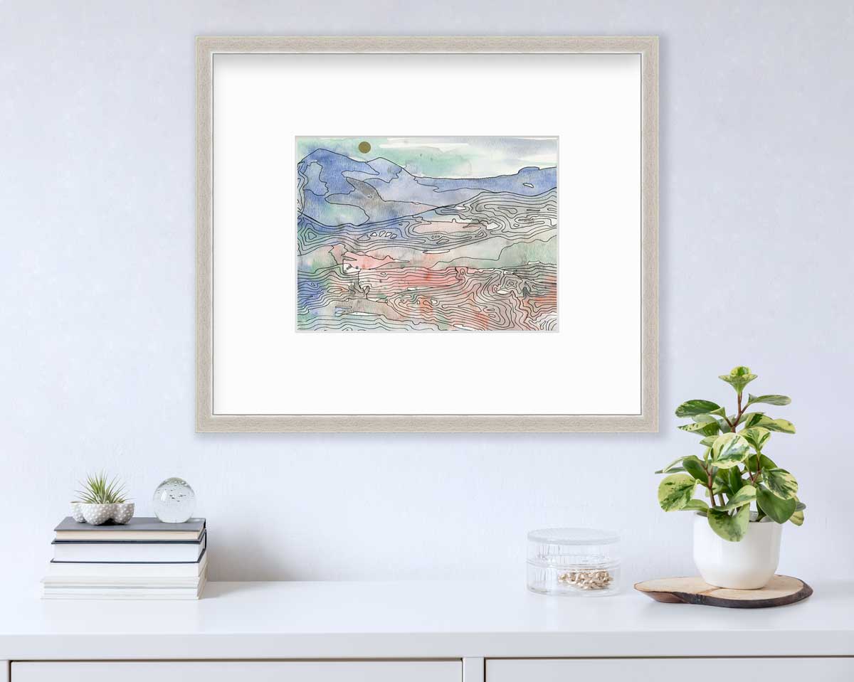 A blue, green and red abstract landscape print in a silver frame with a mat hangs on a white wall above a white surface with a stack of books, a clear round container with gold accessories and a potted plant.