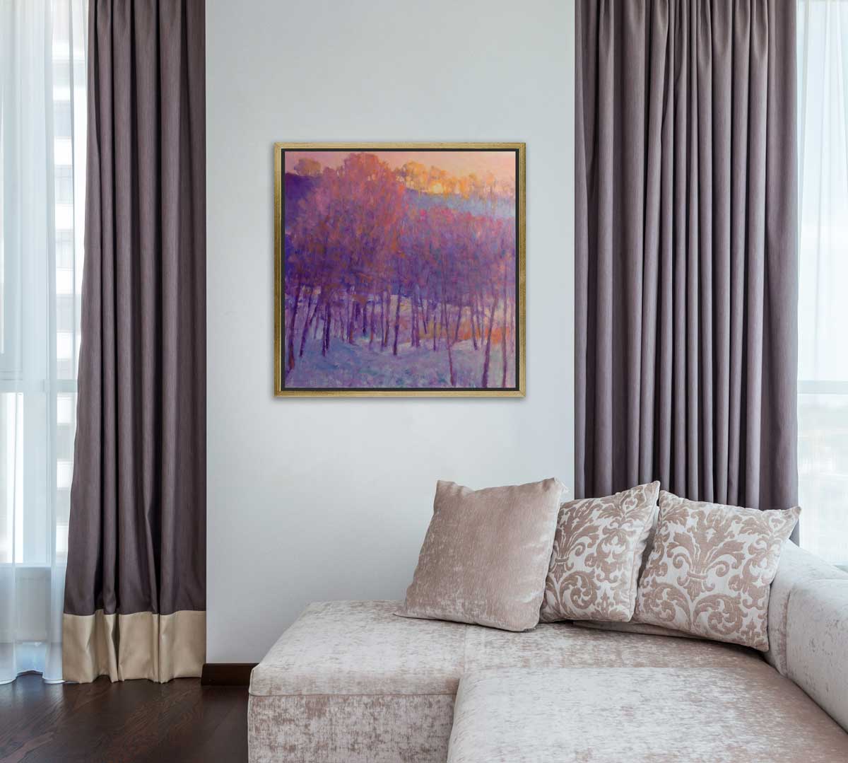 A pink and purple abstract tree landscape print in a gold floater frame hangs on a white wall in between two windows with light mauve curtains and above a pink crushed velvet sofa with pillows.