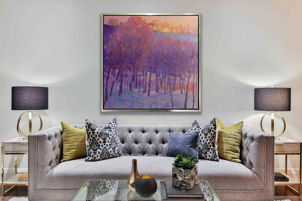 A pink and purple abstract tree landscape in a silver floater frame on a white wall hangs above a grey tufted sofa with pillows, a square glass coffee table with accessories and two side tables with lamps.
