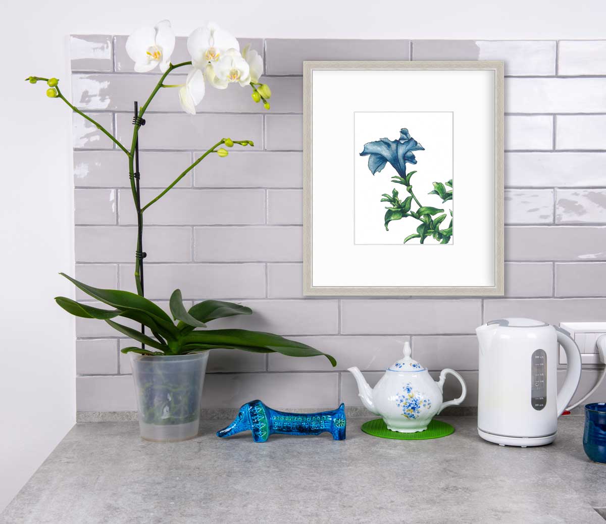 A blue and green flower print in a white frame with a mat hangs on a white tile wall above a grey counter with a potted white orchid, a blue dog figurine, a blue and white teapot and a white water pitcher.