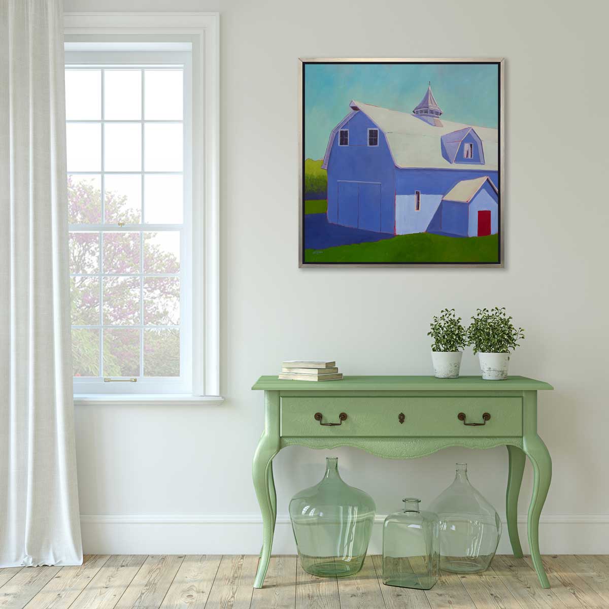 A light blue, lavender and beige contemporary barn print in a silver floater frame hangs on a white wall above a green console table with a stack of books, two potted plants and three light green blown glass vases.