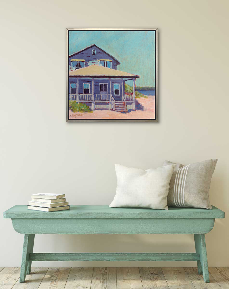 A teal, purple, beige and rose contemporary beach house print in a silver floater frame hangs on a white wall above a teal bench with white and tan pillows and a stack of books.