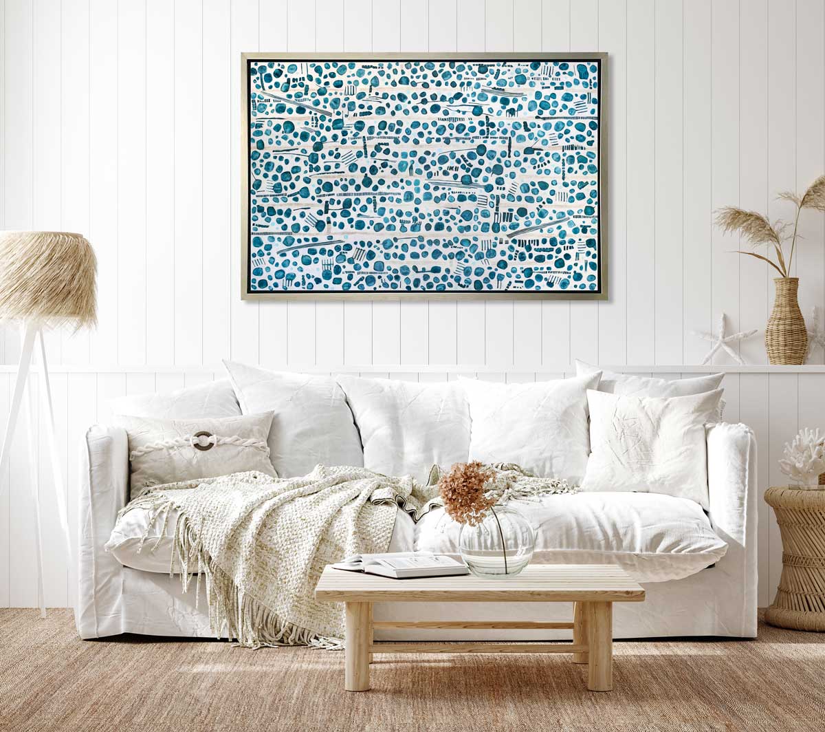A dark teal, white and beige abstract print in a silver floater frame hangs on a white wall above a white sofa with pillows, a natural wood coffee table and a tripod floor lamp.