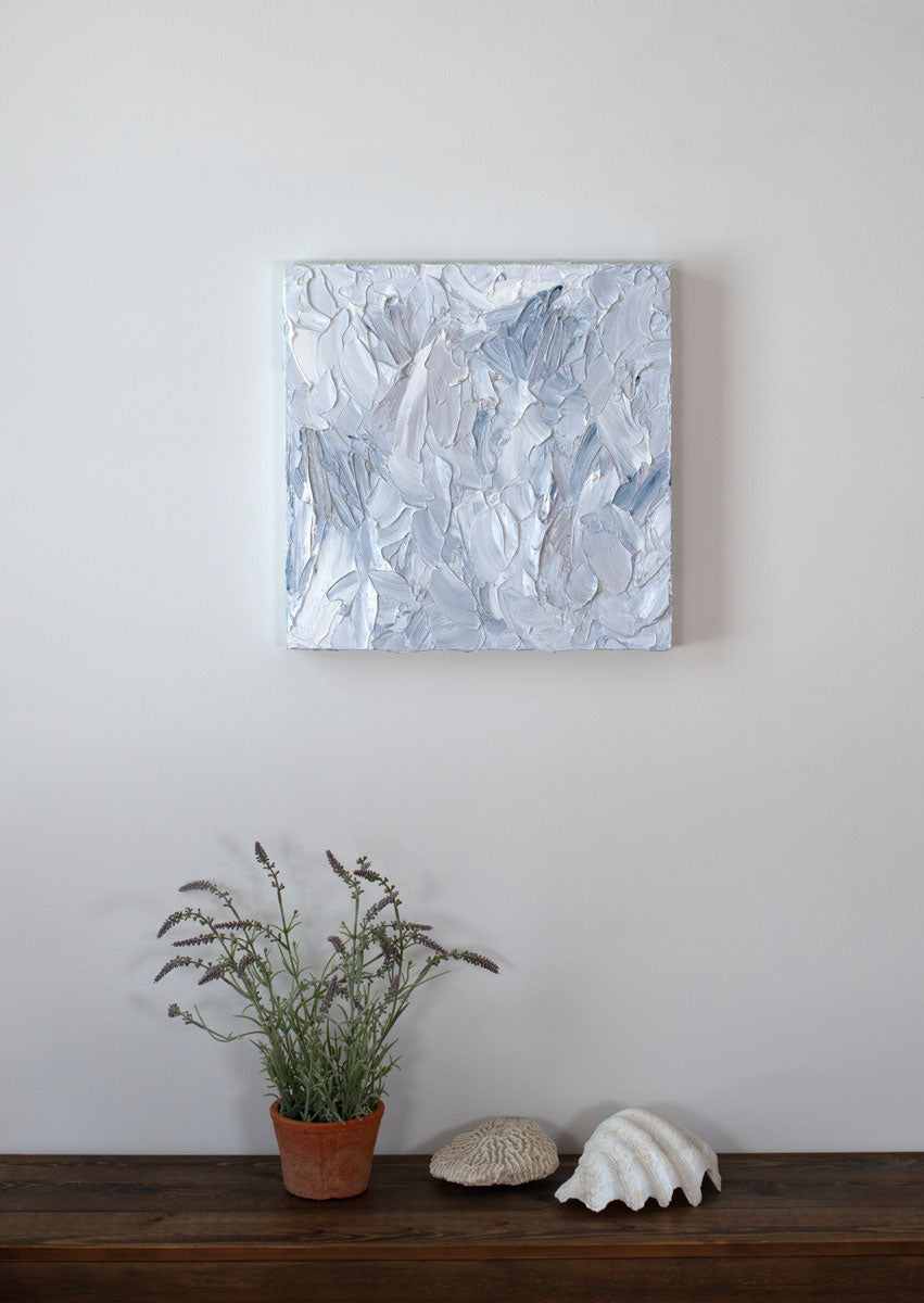 A painting is hung above a shelf. It is decorated in white, blue and grey paint applied in a thick impasto to its wooden surface. Wired and ready to hang.