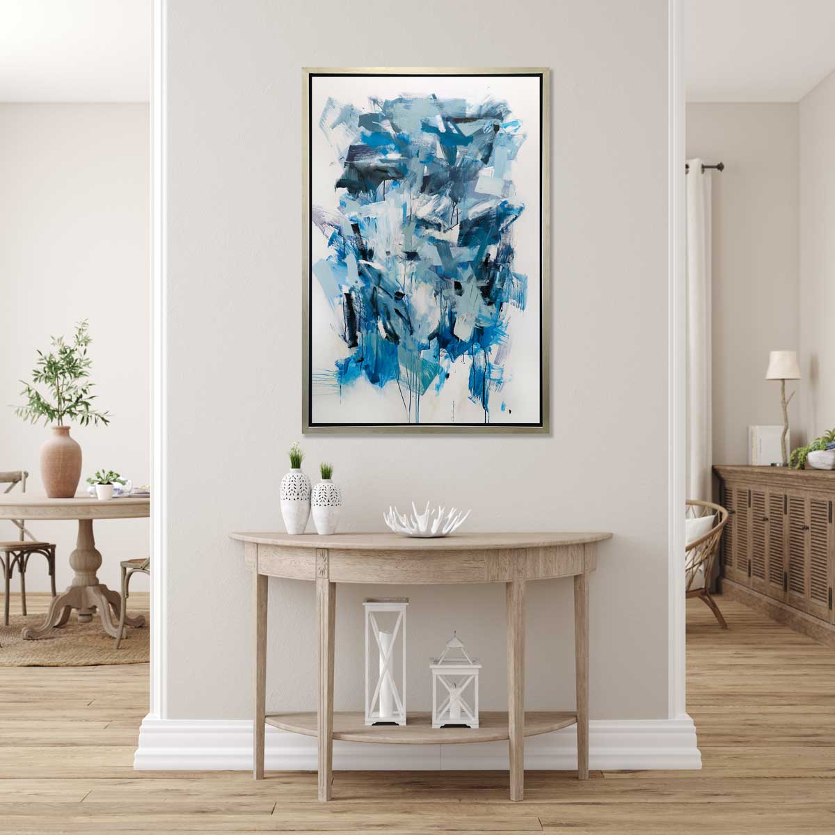 A blue, teal and white abstract print in a silver floater frame hangs on a beige wall above a natural wood half-circle console table with two white vases, a small white decorative bowl and two white lanterns.