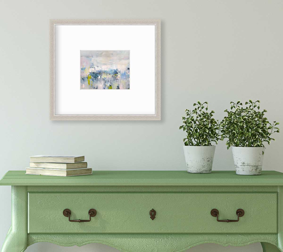 A pink and blue abstract print in a silver frame with a mat hangs on a white wall above a green console table with a stack of books and two potted plants.