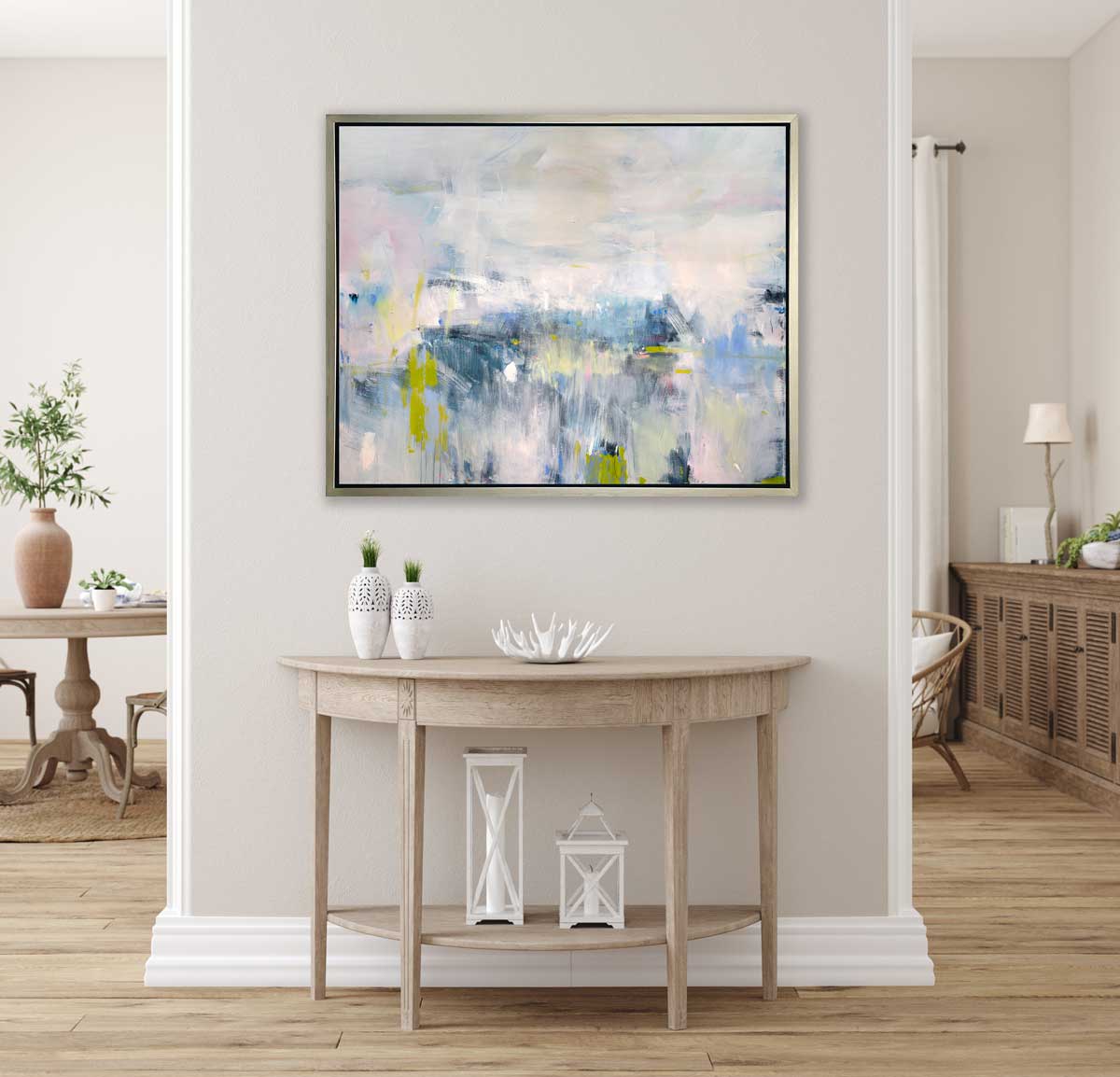 A pale pink, blue and white abstract print in a silver floater frame hangs on a white wall above a natural wood half-circle console table with two white vases, a white decorative bowl and two white lanterns.