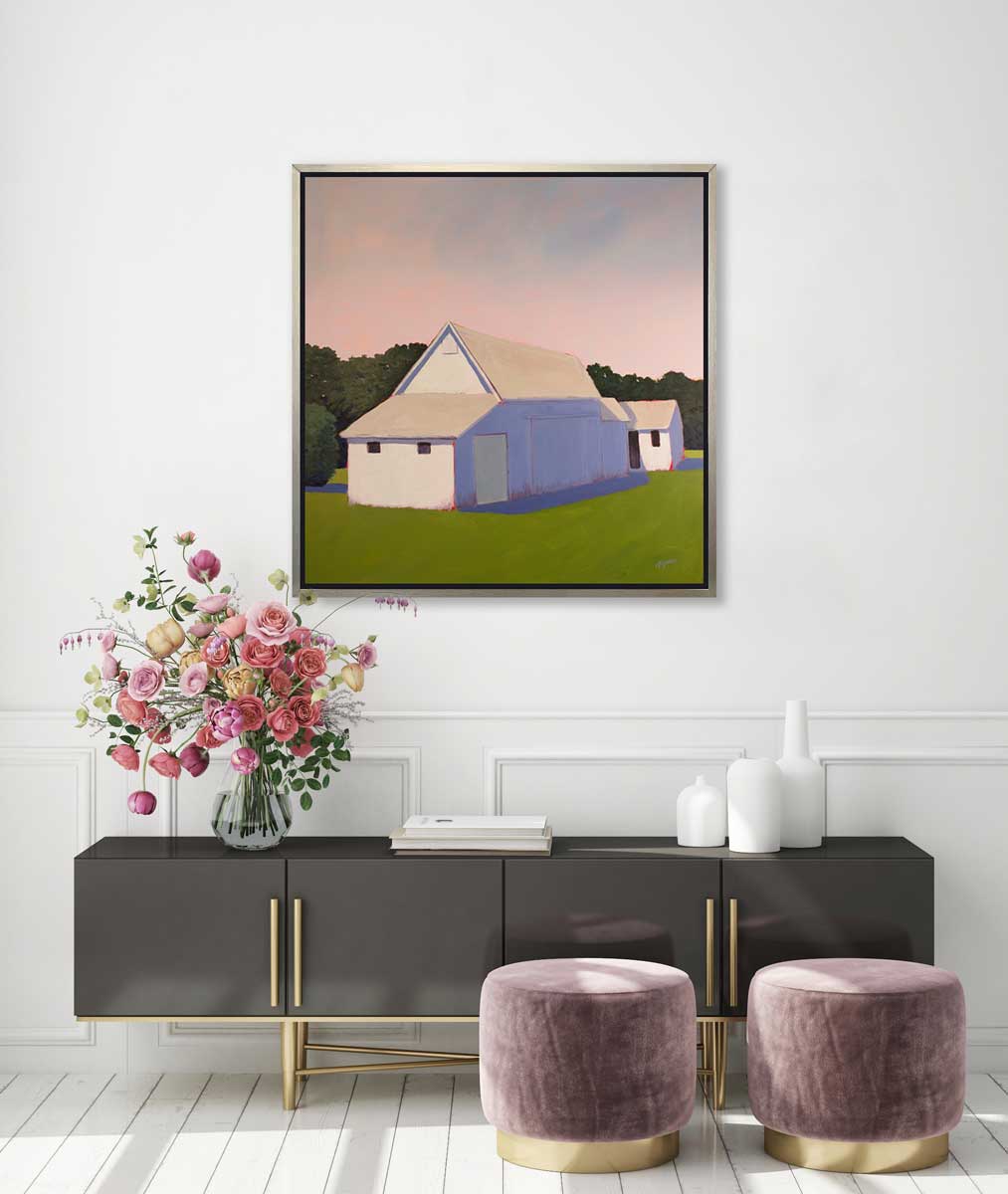 A light pink, lavender and green contemporary barn print in a silver floater frame hangs on a white wall above a black credenza with a clear vase with a colorful floral arrangement, a stack of books, white vases and two mauve ottomans.
