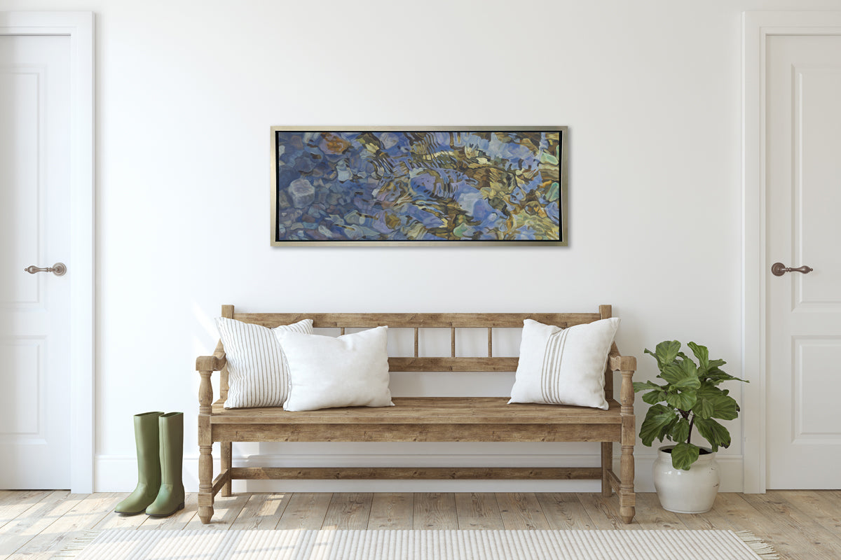 A brown, dark green, white and light blue abstract river rocks in water print in a silver floater frame hangs on a white wall above a natural wood bench with white pillows, a pair of green rain boots and a small potted floor plant.