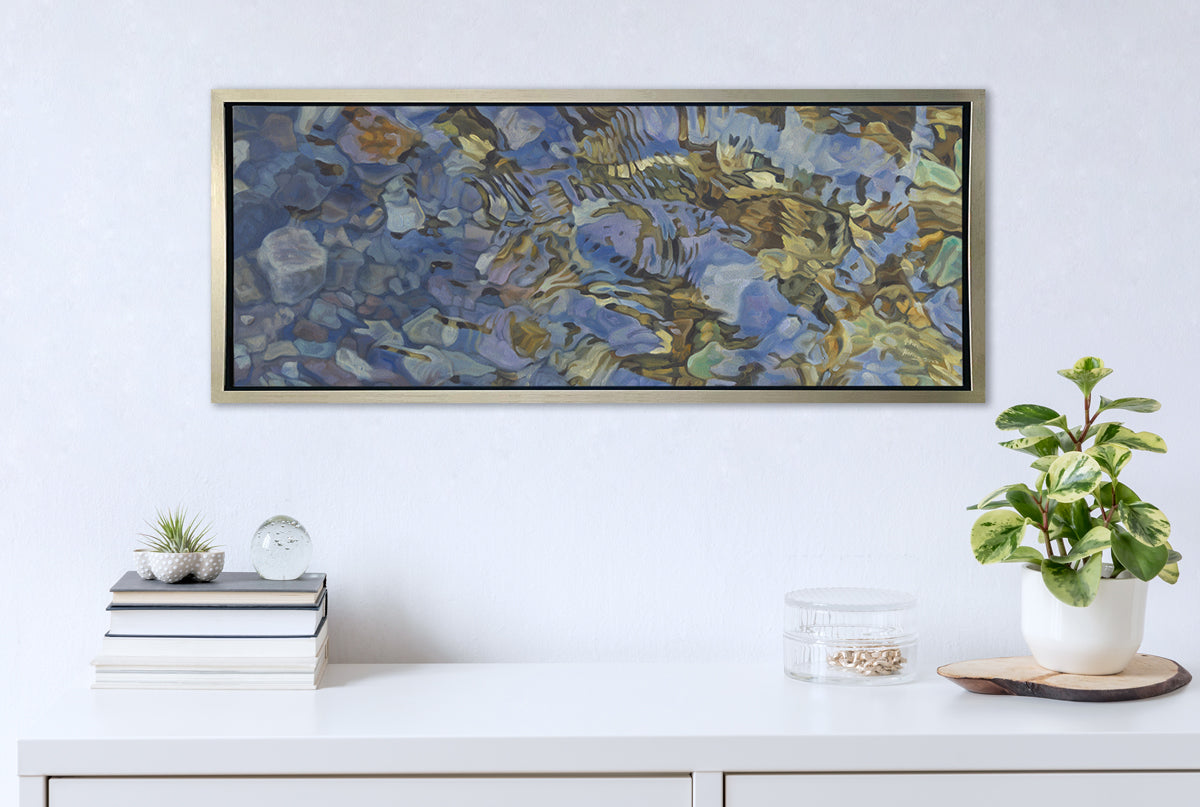 A brown, dark green, white and light blue abstract river rocks in water print in a silver floater frame hangs on a white wall above a white surface with a stack of books, a clear round container with gold accessories and a potted plant.
