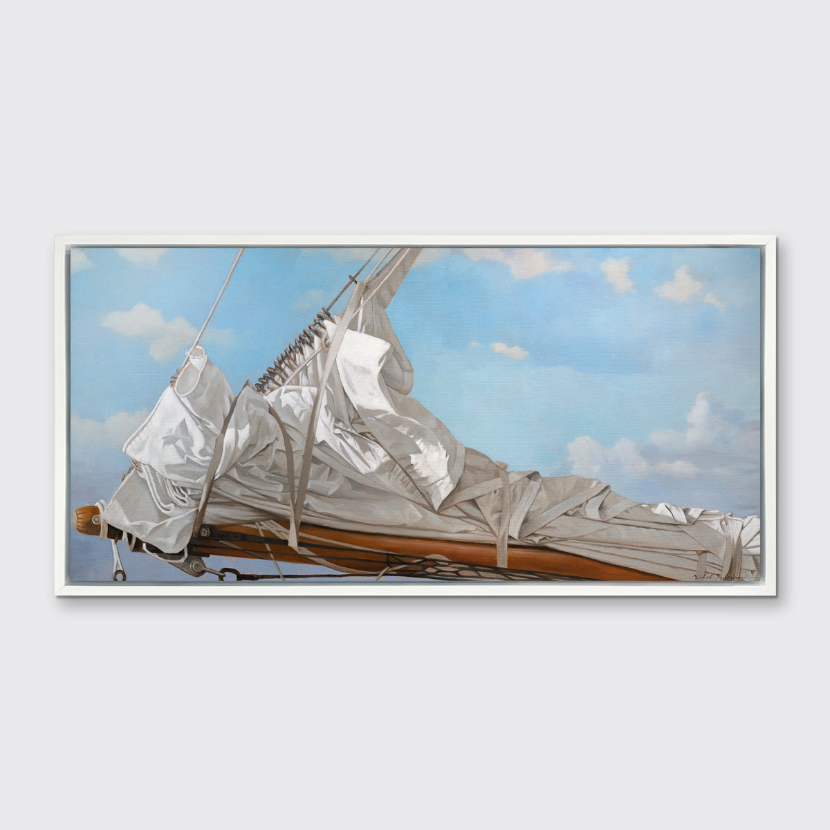 A nautical print of a white sail that has been put down on the bow of a boat against a blue sky with white clouds in a white floater frame hangs on a white wall.