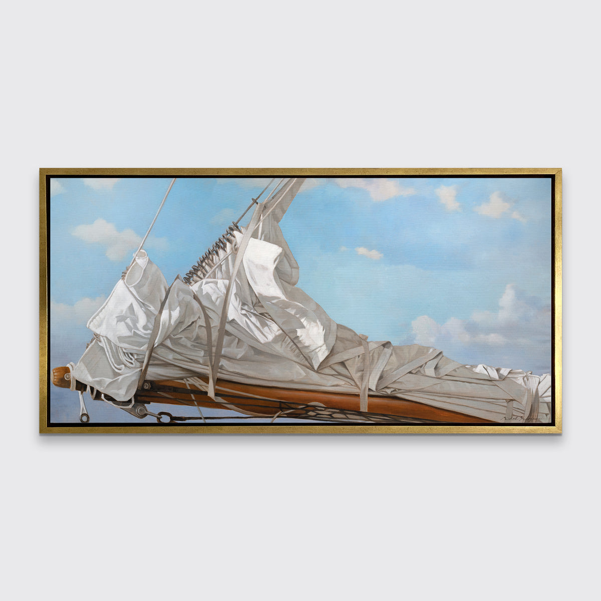 A nautical print of a white sail that has been put down on the bow of a boat against a blue sky with white clouds in a gold floater frame hangs on a white wall.