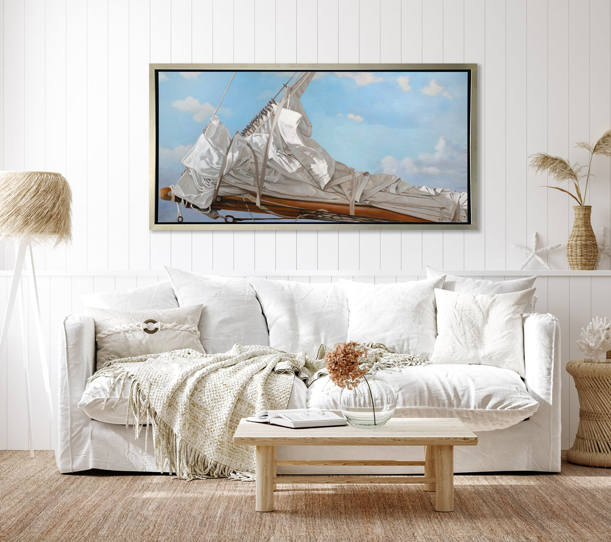 A nautical print of a white sail that has been put down on the bow of a boat against a blue sky with white clouds in a silver floater frame hangs on a white wall above a white sofa with pillows, a natural wood coffee table with an open book and a glass bowl vase with flowers and a tall tripod floor lamp.