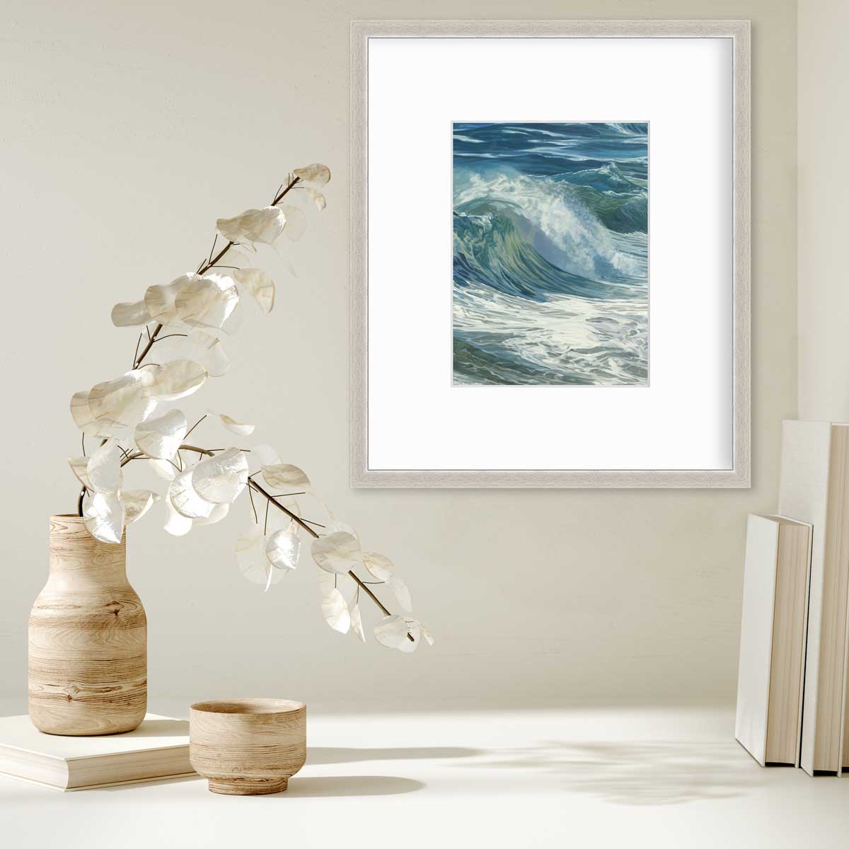 A blue, teal, white and light green abstract water wave print in a silver frame with a mat hangs on a white wall above a stack of books and a wooden vase with white leaf stems.