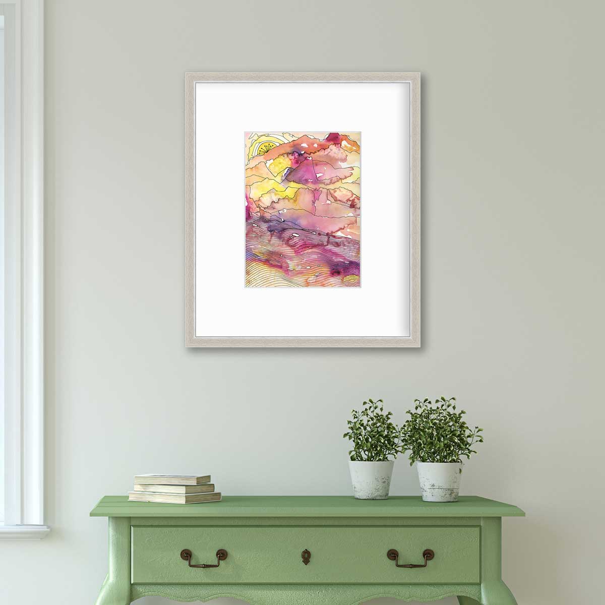 A yellow, pink, orange and black abstract landscape print in a silver frame with a mat hangs on a white wall above a green console table with a stack of books and two potted plants.