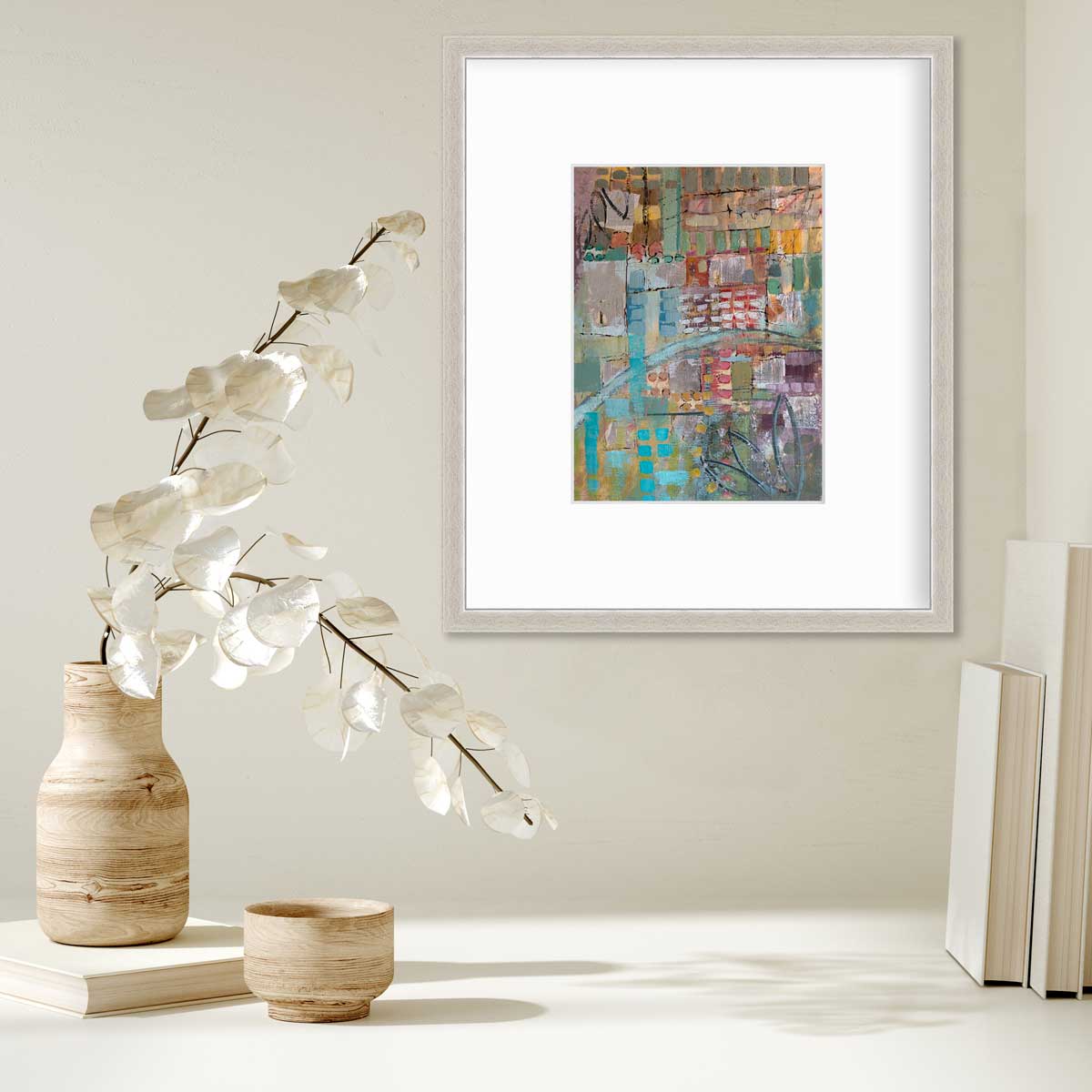 A multicolored abstract print in a silver frame with a mat hangs on a white wall above a stack of books and a wooden vase with white leaf stems.